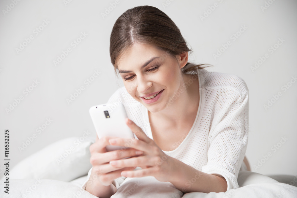Brunette smiling while using smartphone on pillow at home Brunette smiling while using smartphone on pillow at home