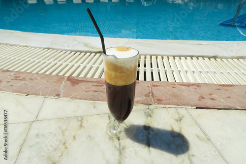 Iced coffee freddo cappuccino by the pool. A glass of cold espresso coffee with steamed milk and a straw by the poolside. photo