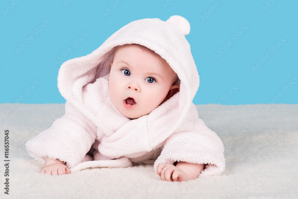 cute baby lying on soft blanket in pink bathrobe with hood and with surprise looks into the camera, isolated on blue cute baby lying on soft blanket in pink bathrobe with hood and with surprise looks into the camera, isolated on blue