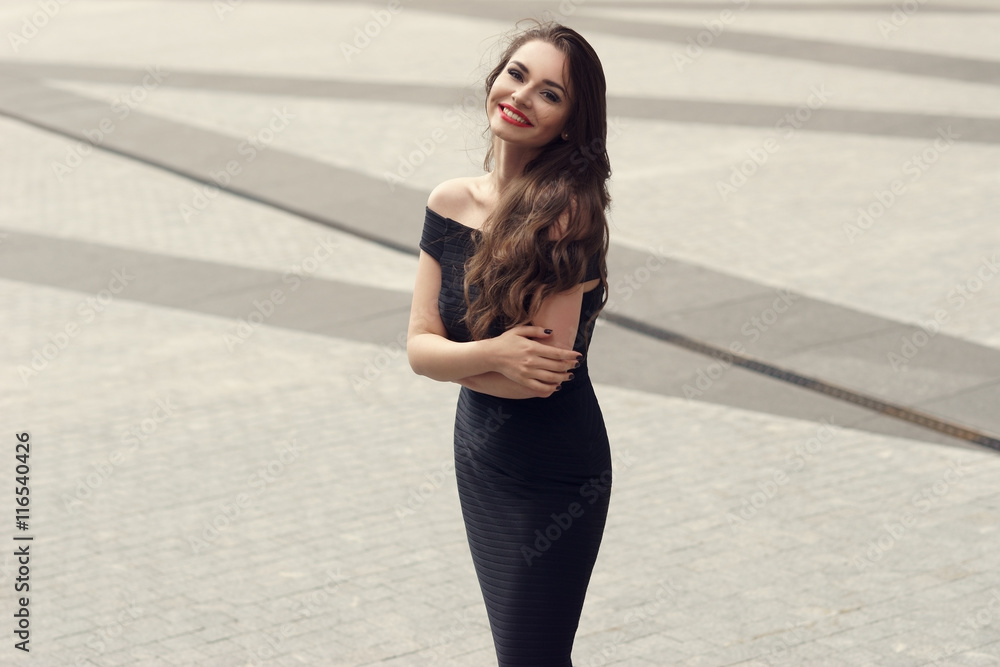 Young pretty woman in sensual black dress standing and posing in city at pavement square