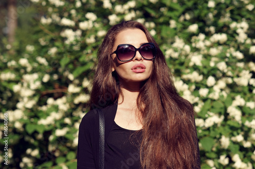 Young beauiful pretty stylish girl in black blouse and sunglasses standing and posing against green bushes with white blooming flowers on a sunny summer day photo