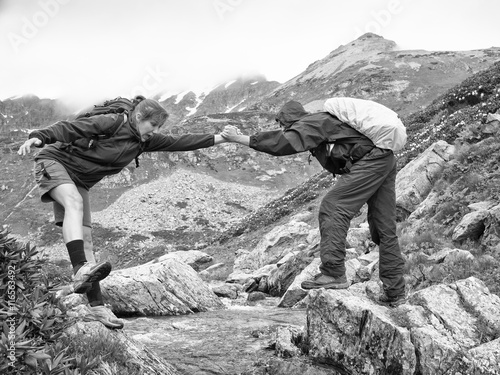 Monochrome image of a young man with a backpack helps frightened woman to overcome the creek on the background of snowy mountains