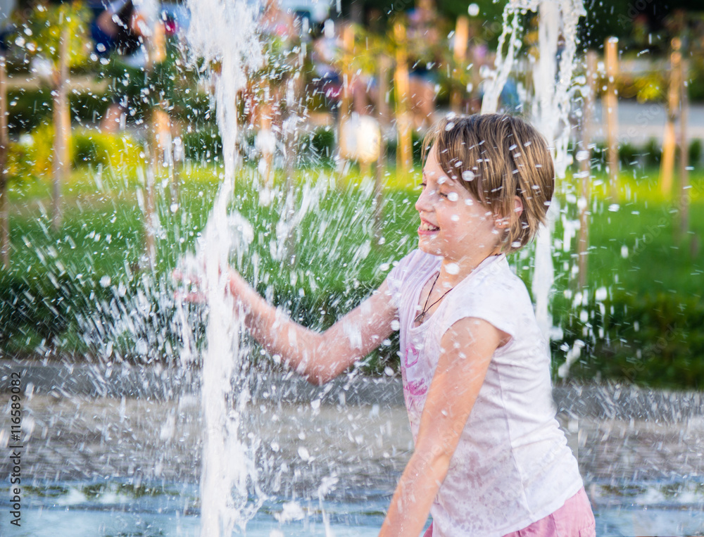 Happy girl having fun between water splashes in fountain . Sunny summer day in the city.