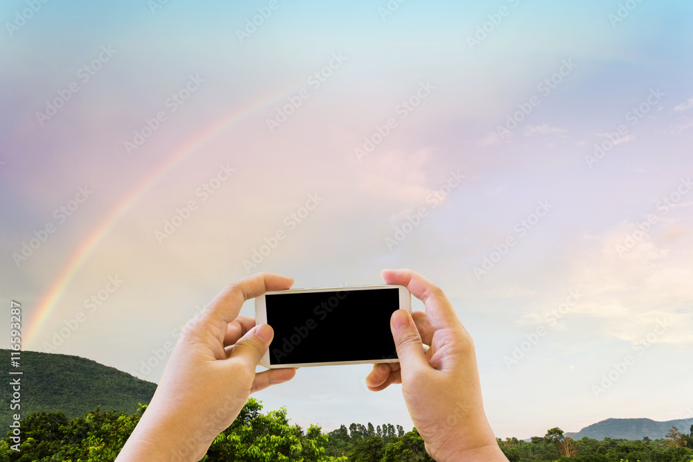 Hand and smartphone or mobile phone taking photo with rainbow