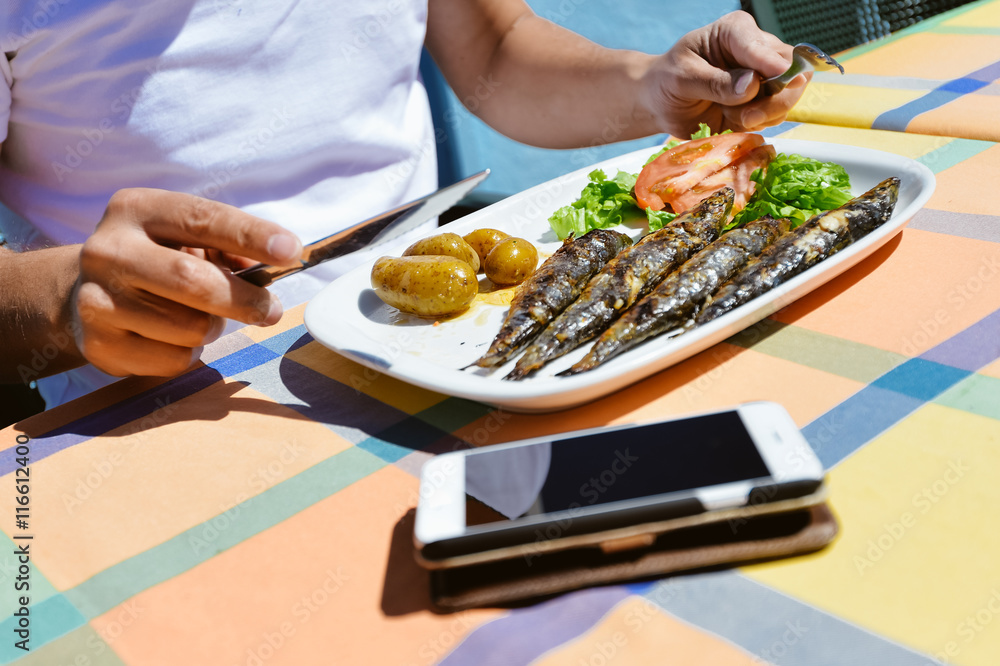 Closeup on mobile phone person eating grilled sardines, restaurant table background. Top side