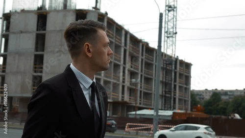 Wallpaper Mural Slow motion. Young serious successful businessman in suit and tie walking near the newly constructed building and looking to unfinished construction project at the background. Middle shot. Torontodigital.ca