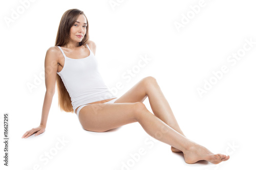 Portrait of beautiful attractive Caucasian young model with long legs sitting against white background and looking at camera. Smiling person posing in tank top and panties. Horizontal studio shot