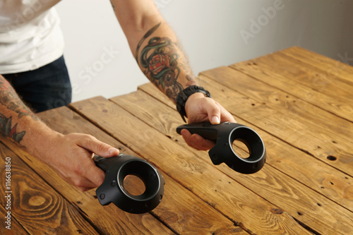 Tattoed hands hold two virtual reality remote controllers above wooden table VR technology presentation photo