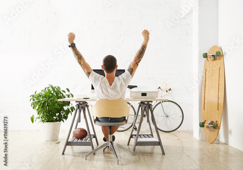Tattooed lucky freelancer in front of his working space, surrounded with his hobby toys longboard, vintage bicycle and green plant, stretching his hand in air while making break photo