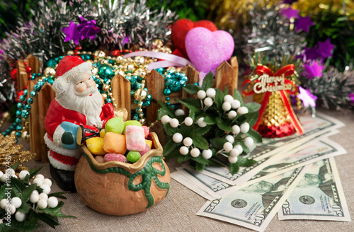 Рождество. Игрушечный Санта Клаус предлагает конфеты. Рядом лежат доллары и сердечки, которые символизируют пожелание богатства и любви. На заднем плане находятся новогодние украшения.