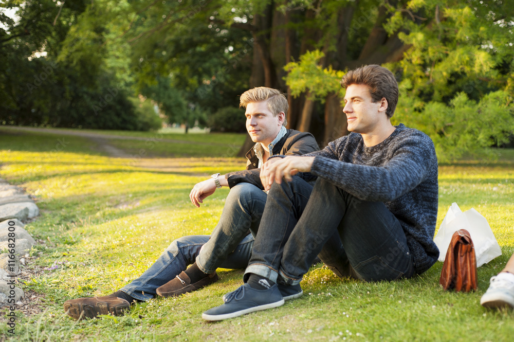 Full length of handsome men looking away while sitting on grassy field in park Full length of handsome men looking away while sitting on grassy field in park