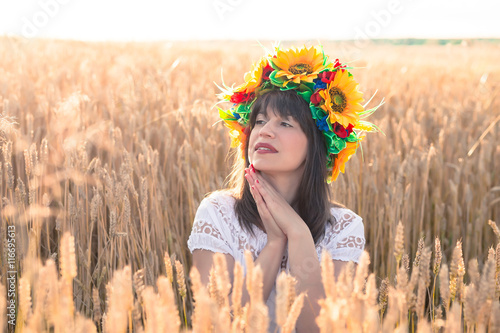 Young woman in a wreath of sunflowers folded his hands near the face and sitting in the wheat