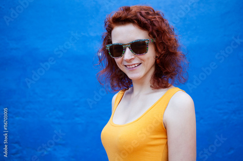 Close up portrait of a ginger girl in sunglasses smiling against blue wall