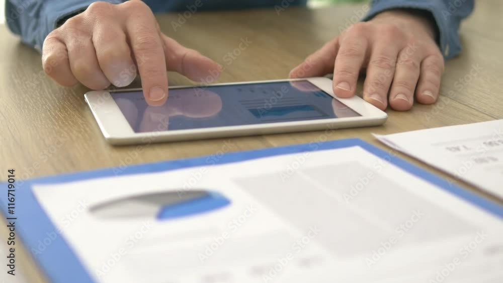Businessman using a touch screen tablet and working at office desk, he is scrolling pages and examining financial charts Businessman using a touch screen tablet and working at office desk, he is scrolling pages and examining financial charts