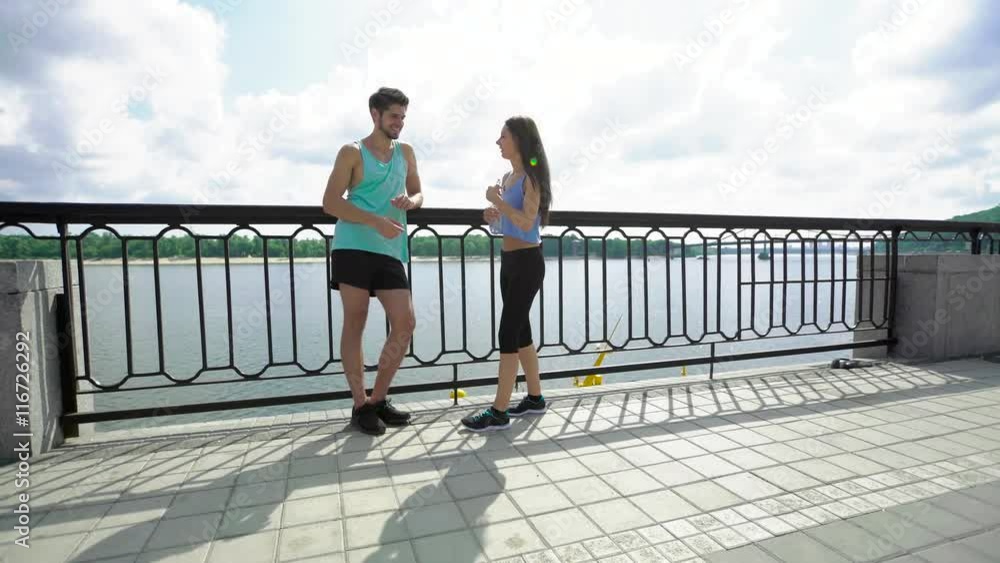 Tired young handsome man resting with bottle of water while smiling woman jogging to him across the bridge in the city Tired young handsome man resting with bottle of water while smiling woman jogging to him across the bridge in the city