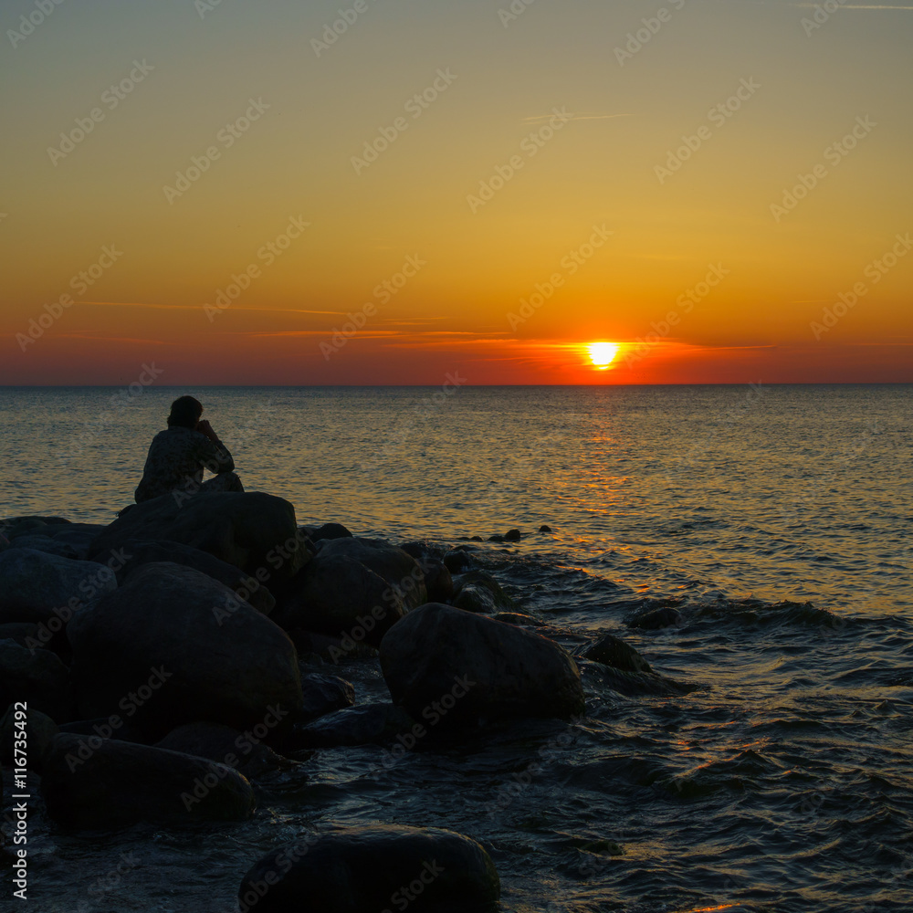 Man sitting on the stones at the seashore and watching beautiful sunset
