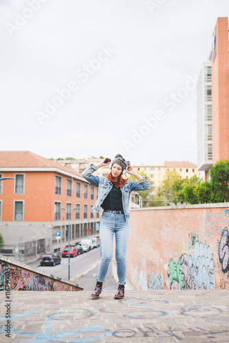 Young handsome caucasian redhead straight hair woman dancing outside in the city, listening music with headphones and smartphone handhold laughing and singing - music, relax, technology concept © Eugenio Marongiu
