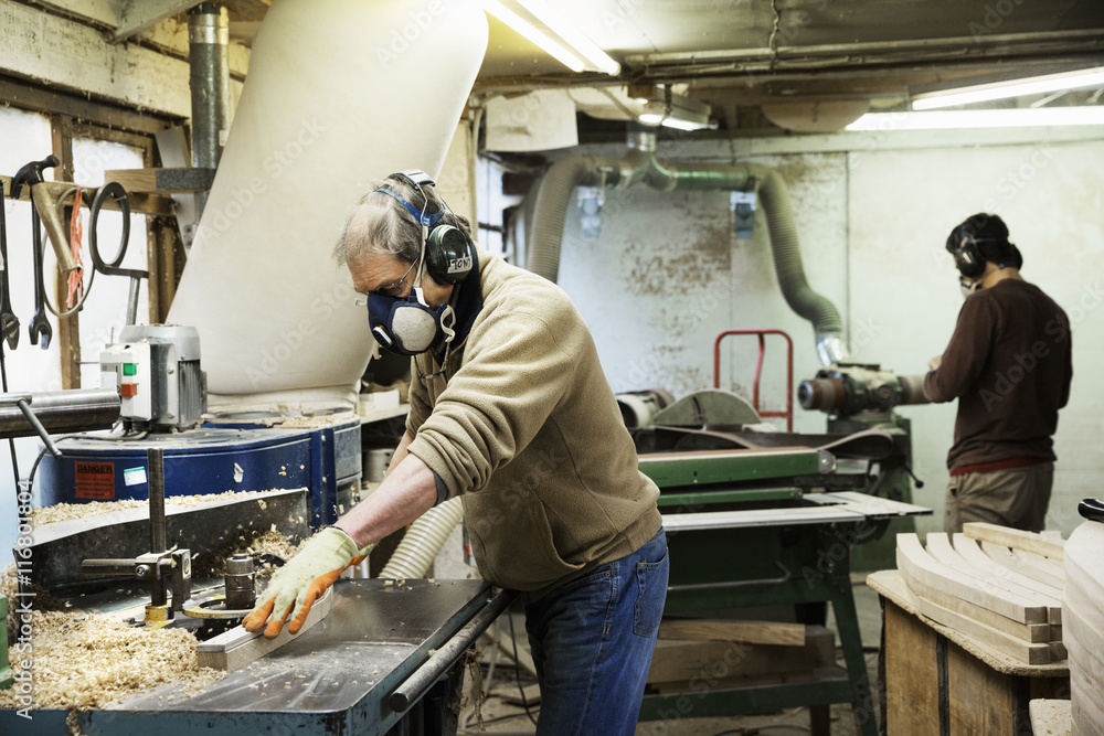 Man standing in a carpentry workshop, wearing a respirator and hearing protector, working on a piece of wood. Man standing in a carpentry workshop, wearing a respirator and hearing protector, working on a piece of wood.