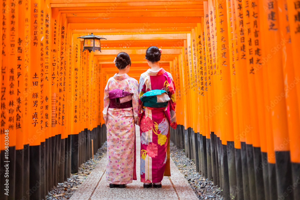 Naklejka premium Dwa gejsze wśród czerwonej drewnianej Tori bramy przy Fushimi Inari świątynią w Kyoto, Japonia. Selektywne focus na kobietach noszących tradycyjne japońskie kimono.