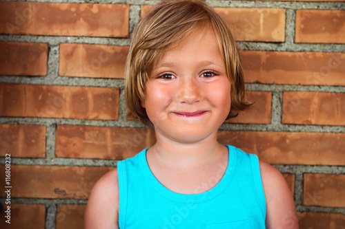 Close up portrait of a cute toddler boy standing against brick wall, wearing blue vest