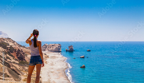 Woman looks on panoramic landscape Petra tou Romiou (The rock of the Greek), Aphrodite's legendary birthplace in Paphos, Cyprus island, Mediterranean Sea. Amazing blue green sea and sunny day.