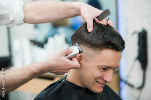 Professional styling. Close up side view of young satisfied man getting haircut by hairdresser with electric razor at barbershop