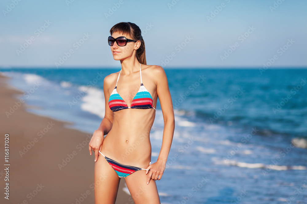 Fashion outdoor portrait young sexy lady having fun and posing in striped bikini and sunglasses on sea background. Sunset light.