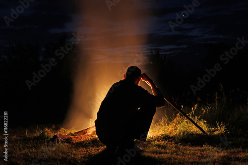 Silhouette of a man with a long stick sitting by the fire at nig