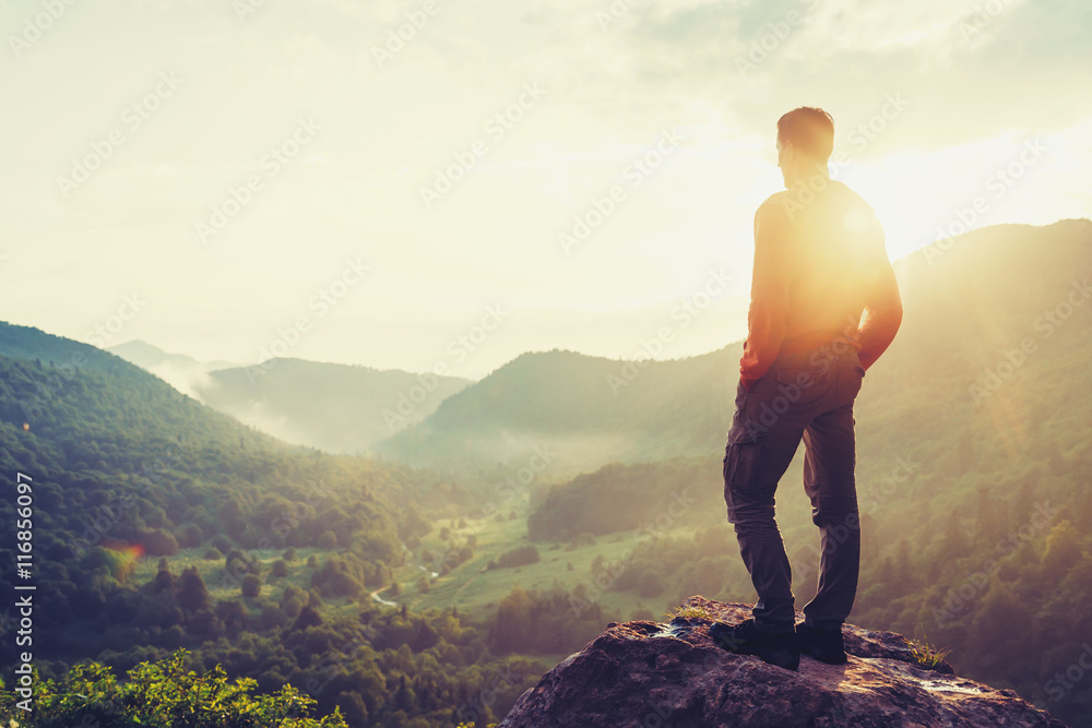 Traveler young man standing in summer mountains at sunset and enjoying view of nature - obrazy, fototapety, plakaty 