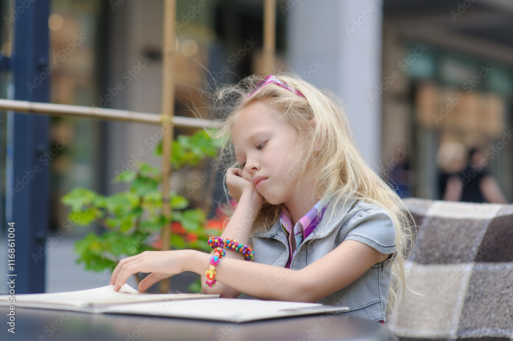 Adorable child reading menu in cafe. Toddler tired girl in beautiful outside cafe choosing meal from menu card. Shop windows in the background. Blond hair fluttering in the wind.