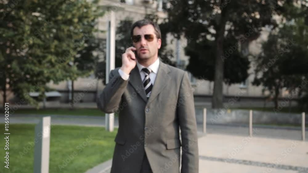 Confident businessman in suit using smartphone outdoors with office building and trees at background. Professional steadycam shot. Confident businessman in suit using smartphone outdoors with office building and trees at background. Professional steadycam shot.