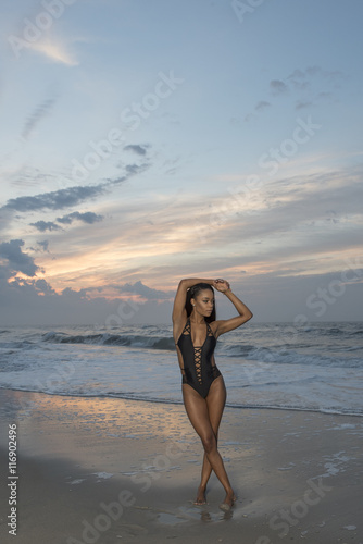 Beautiful African American female model posing on beach in swimsuit at sunrise (sun rising behind her over ocean)