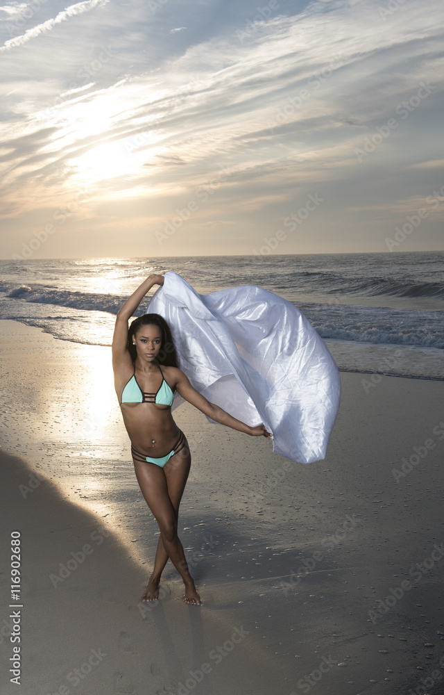 Foto Stock Beautiful African American female model posing on beach in  swimsuit at sunrise (sun rising behind her over ocean) - turquoise colored  bikini - with white stain fabric | Adobe Stock