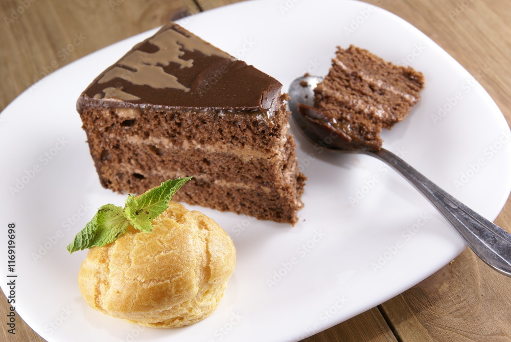 Dessert on a plate on a rustic wooden table: profiterole, decorated with mint leaves and a slice of chocolate cake Dessert on a plate on a rustic wooden table: profiterole, decorated with mint leaves and a slice of chocolate cake