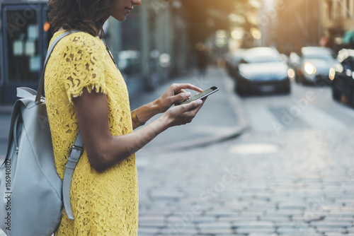 Attractive young girl walking in the city and using an app on smartphone, side view of hipster girl traveling and sending message via her cellphone, sunset in the background