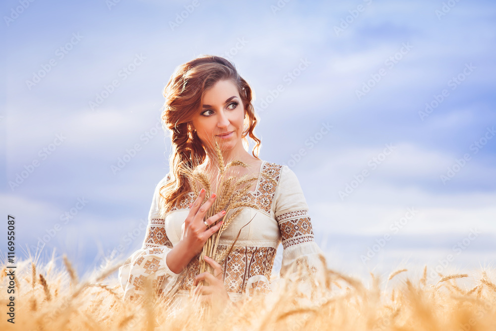 Pretty woman dressed in embroidered gown with tress standing among of wheat field and huging a sheaf of wheat Pretty woman dressed in embroidered gown with tress standing among of wheat field and huging a sheaf of wheat