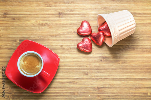 Heart shaped sweets wrapped in a bright red foil lying on wooden texture with  cup of coffe near it. Background for romantic themes.