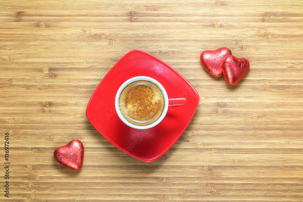Heart shaped sweets wrapped in a bright red foil lying on wooden texture with cup of coffe near it. Background for romantic themes. Heart shaped sweets wrapped in a bright red foil lying on wooden texture with cup of coffe near it. Background for romantic themes.