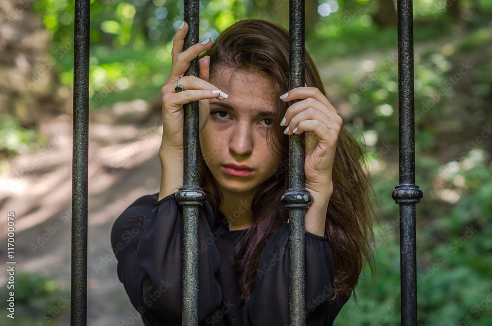 Obraz premium Young charming girl the teenager with long hair sitting behind bars in prison prisoner in a medieval jail with sad, pleading eyes of mercy