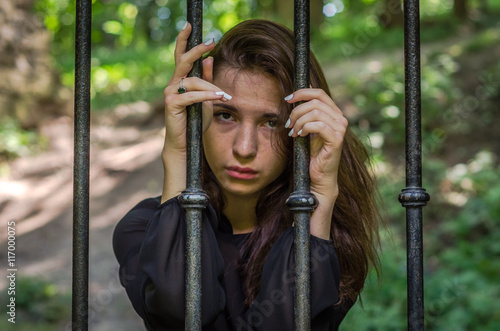 Young charming girl the teenager with long hair sitting behind bars in prison prisoner in a medieval jail with sad, pleading eyes of mercy