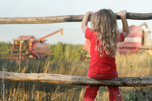 Sitting girl observing farm field with red working combine harvester