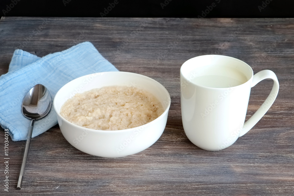 Traditional simple breakfast served on a wooden table.Oatmeal porridge in a white porcelain bowl and a mug of milk. Traditional simple breakfast served on a wooden table.Oatmeal porridge in a white porcelain bowl and a mug of milk.