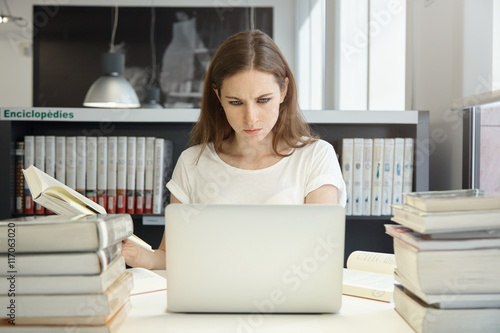 Portrait of female student sitting at the bookshelf and using laptop in the library, reading books, writing thesis with serious face expression. Young Caucasian woman working on her home assignment photo