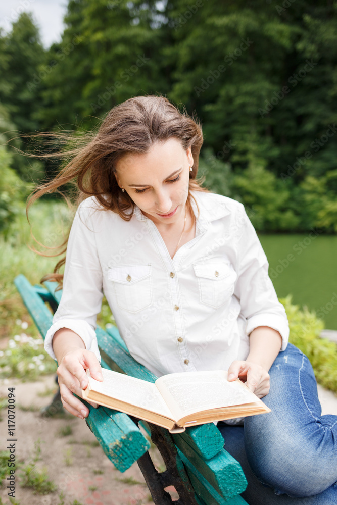 Obraz premium Young woman sitting near pond and reading book