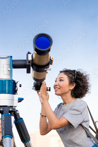 Jeune femme souriante regardant vers le ciel à travers le télescope astronomique Poster Mural XXL