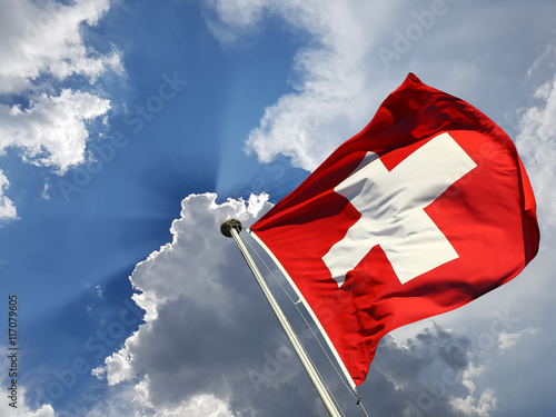 Switzerland national day background: Swiss flag against blue sky with white clouds and sun  rays beaming behind it