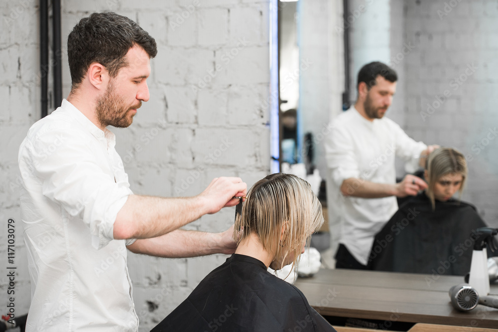 Fototapeta premium hairdresser cuts hair with scissors on crown of handsome satisfied client in professional hairdressing salon
