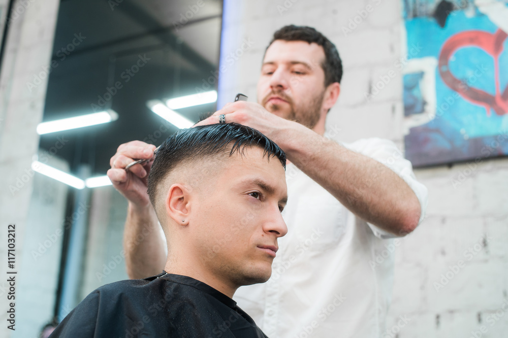 Fototapeta premium Professional styling. Close up side view of young man getting haircut by hairdresser with electric razor at barbershop
