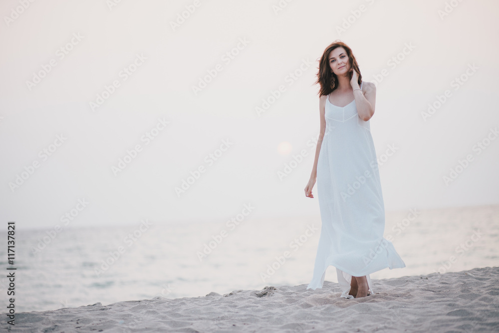 Young beautiful woman in a white dress walking on an empty beach near ocean Young beautiful woman in a white dress walking on an empty beach near ocean