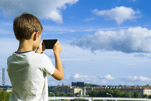 Boy with mobile phone. Child taking photo his smartphone. Beautiful sky and city background. Back view. Technology concept